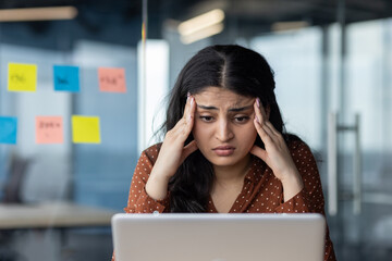 Young hispanic businesswoman stressed in office, working on laptop. Frustrated, overwhelmed woman...