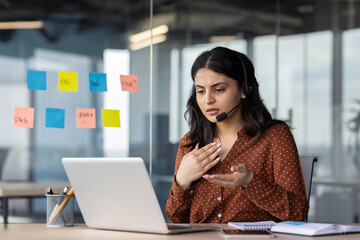 Young Hispanic business woman using laptop and headset during virtual meeting. She looks focused as she discusses solutions in modern office. Concepts of communication, technology, work, business.