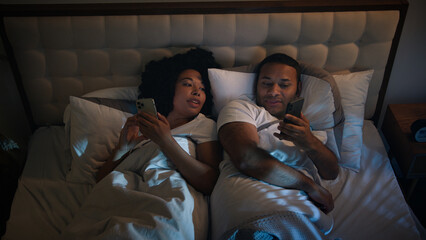 African American couple in bed at night late time in game bedroom relaxing using two phones chatting in mobile app scrolling social media smartphones woman man wife husband talking discuss news family