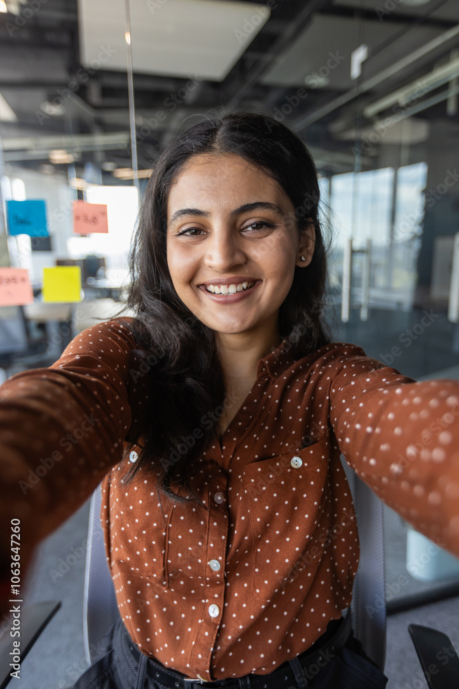 Wall mural Young hispanic business woman in modern office setting using phone for video call. She is smiling, showcasing connectivity and communication in professional environment.