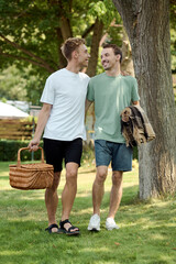 Two happy men walk in a sunny park, carrying a picnic basket and enjoying each others company.