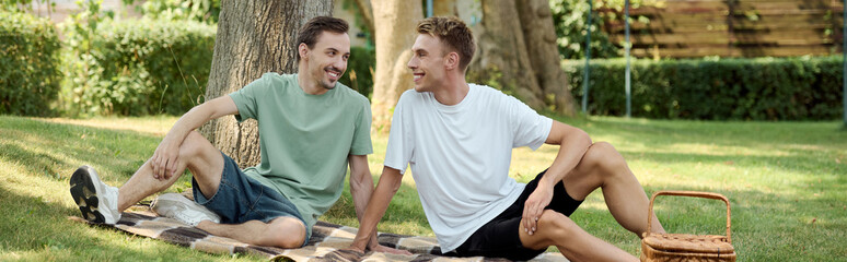 Two joyful men share a picnic on a sunny day, surrounded by nature and laughter.