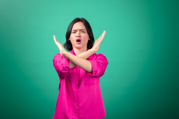 An Asian woman in a pink shirt stands against a green background, crossing her arms in an "X" shape in front of her, with a serious expression. The gesture emphasizes a strong "no" or disapproval.