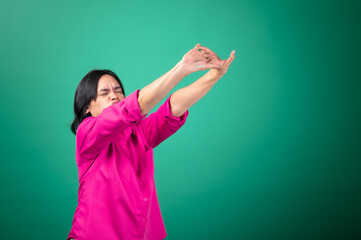 Asian woman in a bright pink shirt against a green background appears to be yawning, with arms...