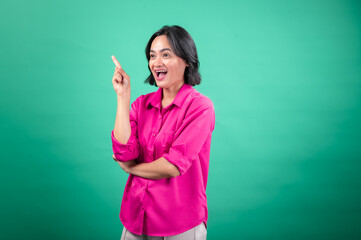 An Asian woman in a vibrant pink shirt stands against a green background, looking cheerful and pointing upwards with one hand, as if indicating a bright idea or something exciting