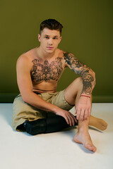 A tattooed young man showcases his prosthetic leg and striking ink in a vibrant studio environment.