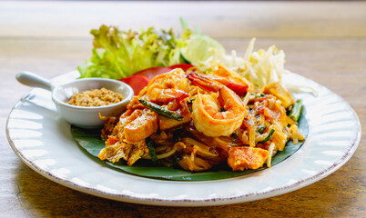 Fried noodle thai style with prawns, pad thai with prawns, bean sprout, garnish peanuts, lemon lime and fresh vegetables on white plate on wooden table. Selective focus.