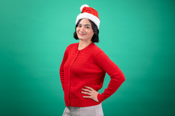 An Asian woman wearing a bright red, buttoned sweater and a festive Santa hat poses with hands on her hips against a green background, smiling confidently in a cheerful holiday-themed setting