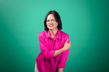 Obraz premium An Asian woman wearing a bright pink shirt against a green background is holding her shoulder with one hand, expressing pain or discomfort, with her face showing a pained expression