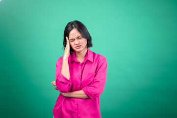 An Asian woman in a bright pink shirt against a green background holds her hand to her head, eyes closed, expressing a feeling of headache or stress.