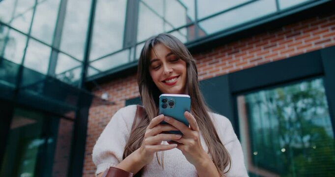 Pretty Caucasian girl standing outside in front of business center. Holding smartphone with both hands. Reading message on screen. Thinking what to reply. Chatting online with someone.