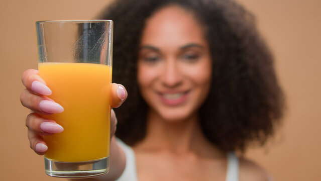 Happy healthy African American woman ethnic girl female model drinking fresh orange juice vitamin drink stretch hand with glass motivate clink clinking suggest to try diet detox health care in studio