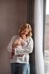 Fototapeta premium Young beautiful mother holds newborn baby while standing at home near window. tender moment with baby dressed in soft pink And mom in light clothes. Natural light, peaceful and intimate atmosphere