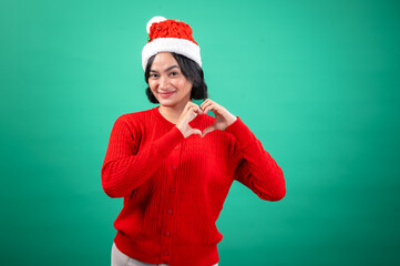 An Asian woman wearing a red sweater and a Santa hat forms a heart shape with her hands, expressing warmth and love. She stands against a green background, adding a festive