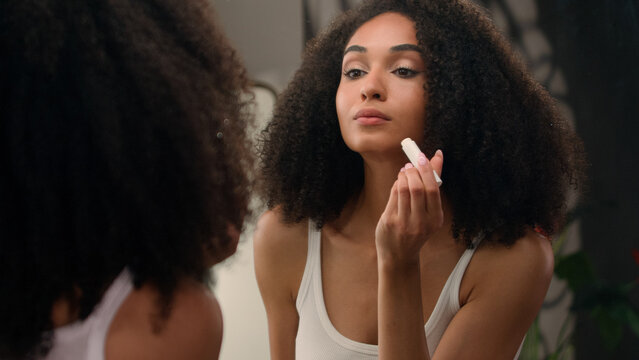 Beautiful African American woman looking in mirror reflection morning beauty routine preparation ethnic girl in bathroom applying moisturizing lip balm lipstick smooth lips organic natural cosmetic