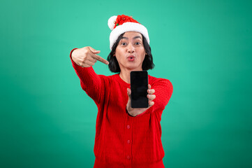 An Asian woman wearing a red sweater and Santa hat stands against a green background, holding a smartphone in front of her with an excited expression and pointing to it