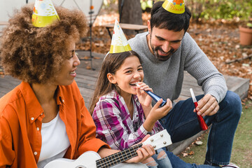 Young mixed-race interracial family of three with daughter celebrating birthday party outdoors,...