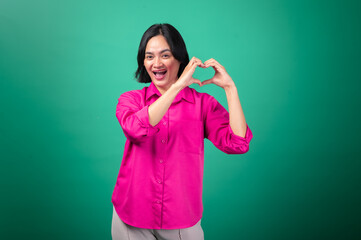 Fototapeta premium An Asian woman in a pink shirt stands against a green background, forming a heart shape with her hands in front of her chest and smiling warmly, conveying love, positivity, and affection.