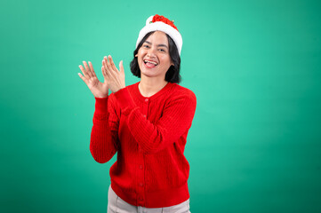 An Asian woman wearing a red sweater and Santa hat claps her hands with a joyful expression, conveying festive energy. She stands against a green background,