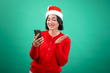 An Asian woman in a Santa hat and red sweater smiles at her phone, gesturing cheerfully with her hand, set against a green background.