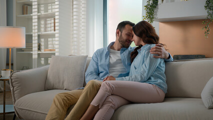 Cheerful beautiful young couple in love hugging cuddling relaxing on couch at modern home touch foreheads cuddle hug together feel love tender bonding affectionate enjoy romantic date wife husband