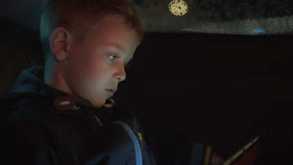 Little boy sitting in the back seat of car and watching video on tablet at night