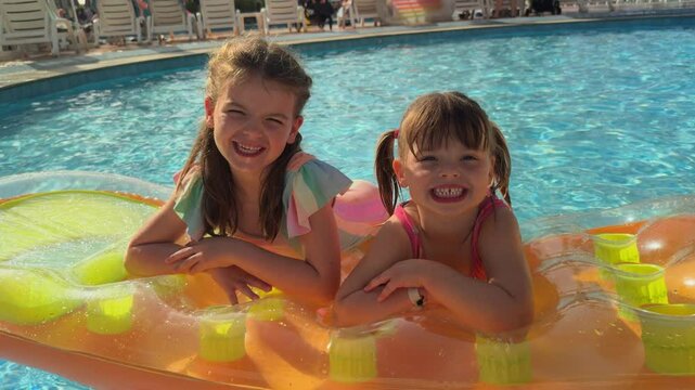 4K: Two little girls smiling at the camera lying on an Inflatable Lilo in the Swimming pool on Vacation Holiday. Stock Video Clip Footage