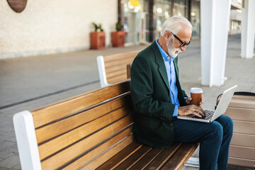 Senior man sits at a train station and enjoys a coffee, working on laptop during the day.