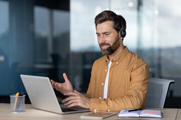 Mature businessman engaged in virtual meeting, wearing headset with laptop in office. Displays...