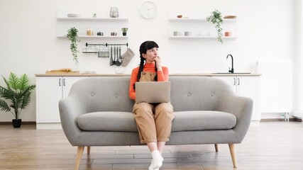 Young woman with dark hair sitting on couch using laptop in bright, contemporary living room. Casual attire and relaxed posture suggest work-from-home or study environment.
