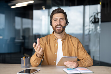 Confident businessman using headset, engaging in virtual meeting. Enthusiastic expression, holding notebook, gesturing during online discussion. Tablet and phone on desk enhance modern office setting