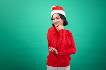 An Asian woman in a red sweater and Santa hat stands against a green background, smiling thoughtfully with one hand resting under her chin, capturing a festive