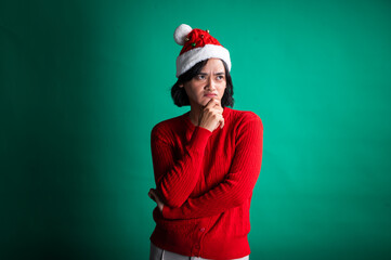 An Asian woman in a red sweater and Santa hat poses with a thoughtful expression, resting her chin on her hand, against a green background, creating a humorous and festive holiday mood