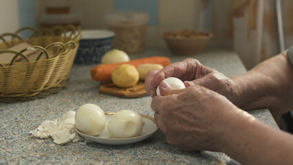 Elderly woman peeling boiled egg while sitting in modest kitchen. Hands close-up