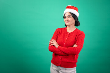 Asian woman wearing a red sweater and festive Santa hat stands confidently with her arms crossed against a green background, exuding a calm holiday spirit in a casual Christmas-themed