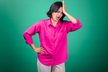 An Asian woman in a bright pink shirt against a green background holds her hand to her head, eyes closed, expressing a feeling of headache or stress.