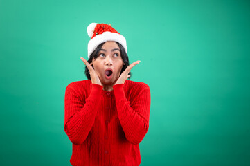 An Asian woman in a red sweater and Santa hat holds her hands to her face with an expression of surprise and wide eyes, standing against a green background. She exudes a festive, playful vibe