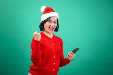 An Asian woman in a Santa hat and red sweater celebrates while looking at her phone, showing excitement and joy, set against a green background.