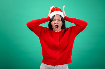 An Asian woman wearing a red sweater and Santa hat holds her hands on her head with a surprised expression, as if shocked or overwhelmed. She stands against a green background