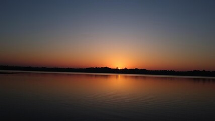A serene landscape at dusk, with the sun setting behind silhouetted trees and reflecting on a calm lake, outdoors, natural