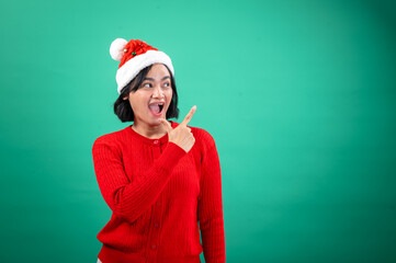 An Asian woman in a red sweater and Santa hat points enthusiastically to the side with an open-mouthed smile, standing against a green background, capturing a festive and cheerful holiday vibe