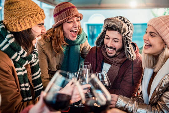 Young people  toasting red wine dining at restaurant terrace - Happy friends group socializing drinking and eating food sitting outside at winery bar table - Winter season, dinner lifestyle concept