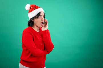 An Asian woman in a red sweater and Santa hat stands against a green background, appearing to shout or call out with her hand cupped around her mouth, expressing excitement and energy