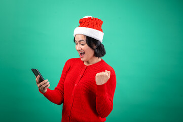 An Asian woman in a Santa hat and red sweater celebrates while looking at her phone, showing excitement and joy, set against a green background.