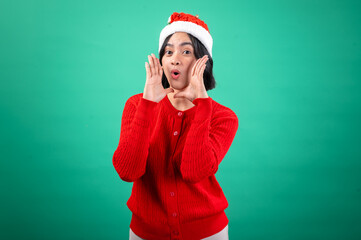 An Asian woman in a red sweater and Santa hat stands against a green background, appearing to shout or call out with her hand cupped around her mouth, expressing excitement and energy