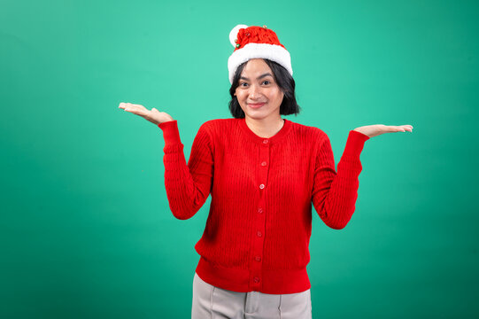 An Asian woman in a red sweater and Santa hat shrugs with both hands raised, expressing confusion or indifference, standing against a green background. Her facial expression is questionin