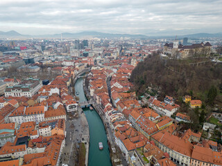 Naklejka premium Panorama of Ljubljana city. Aerial view above Ljubljanica river with river cruise boats towards city center. Historical town with old architecture and the castle on the hill. Cloudy day in the winter.