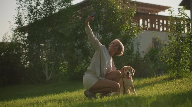 Woman taking selfie with her dog focused on something else outdoors in lush green garden under sunny weather, with building and trees in background, dog is sitting, panting, while owner smiles
