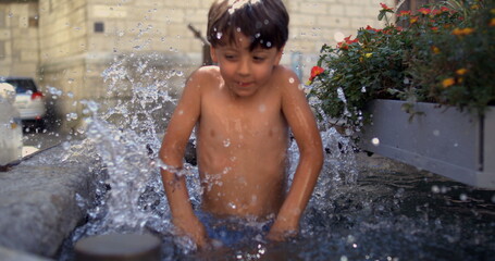 Young boy jumping into water, splashing playfully in a fountain, surrounded by flowers, in a...