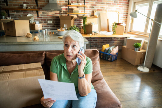 Senior woman making a phone call while reviewing paperwork in living room with moving boxes